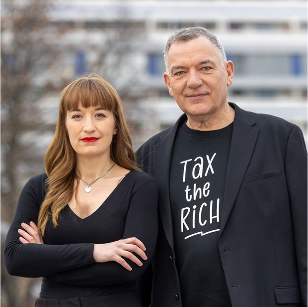 Jan van Aken steht neben Heidi Reichinnek, der Co-Spitzenkandidatin von Die Linke für die Bundestagswahl 2025. Beide tragen schwarze Kleidung und blicken entschlossen in die Kamera. Jan van Aken trägt ein schwarzes T-Shirt mit der weißen Aufschrift „TAX THE RICH“ unter einem schwarzen Blazer. Der Hintergrund zeigt eine städtische Kulisse mit modernen Gebäuden und kahlen Bäumen.