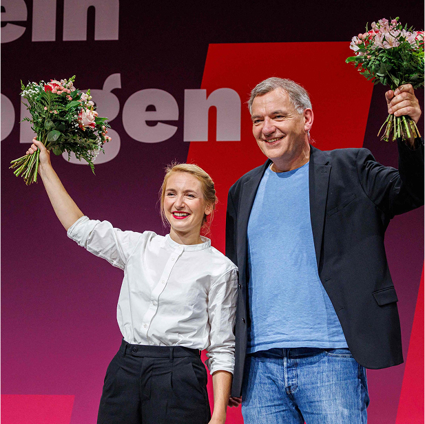 Jan van Aken steht auf der Bühne neben Ines Schwerdtner, der Co-Vorsitzenden von Die Linke. Beide halten strahlend einen Blumenstrauß in die Luft. Das Bild wurde auf dem Parteitag von Die Linke aufgenommen, bei dem sie gemeinsam als neue Vorsitzende gewählt wurden. Im Hintergrund ist ein großes rotes Banner mit weißem Schriftzug zu sehen, das zur Parteikampagne gehört.