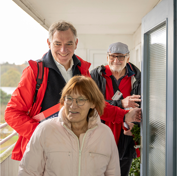 Jan van Aken nimmt an einer Haustüraktion (Haustürgespräche) mit Mitgliedern von Die Linke teil. Er trägt eine rote Jacke und hält Wahlkampfmaterial in der Hand. Neben ihm steht ein älterer Genosse mit Brille und Mütze, ebenfalls in einer roten Jacke. Gemeinsam sprechen sie mit einer älteren Frau, die die Tür geöffnet hat. Die Szene spielt sich in einem Wohnhausflur ab und zeigt den direkten Kontakt mit Wähler:innen.