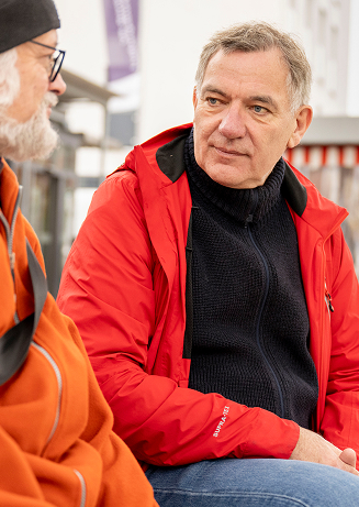 Jan van Aken ist in einer Nahaufnahme zu sehen, wie er aufmerksam mit einem älteren Mann mit weißem Bart und Brille spricht. Er trägt eine rote Jacke über einem schwarzen Rollkragenpullover, was auf eine Wahlkampfaktion im Freien hindeutet. Der Hintergrund ist leicht unscharf und zeigt Marktstände mit rot-weißen Markisen, was auf eine belebte Umgebung hindeutet. Im unteren linken Bereich des Bildes steht in großen roten Buchstaben auf weißem Hintergrund „Jan van Aken“. Die Szene vermittelt einen direkten Austausch mit den Bürger:innen im Wahlkampf.